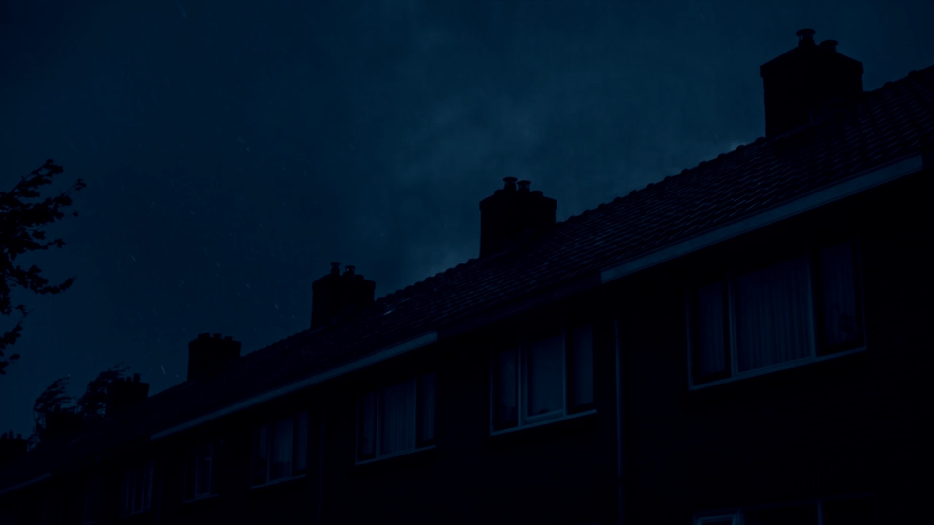 Terraced rooftops under a dark storm sky at night, with rain driving across the chimneys