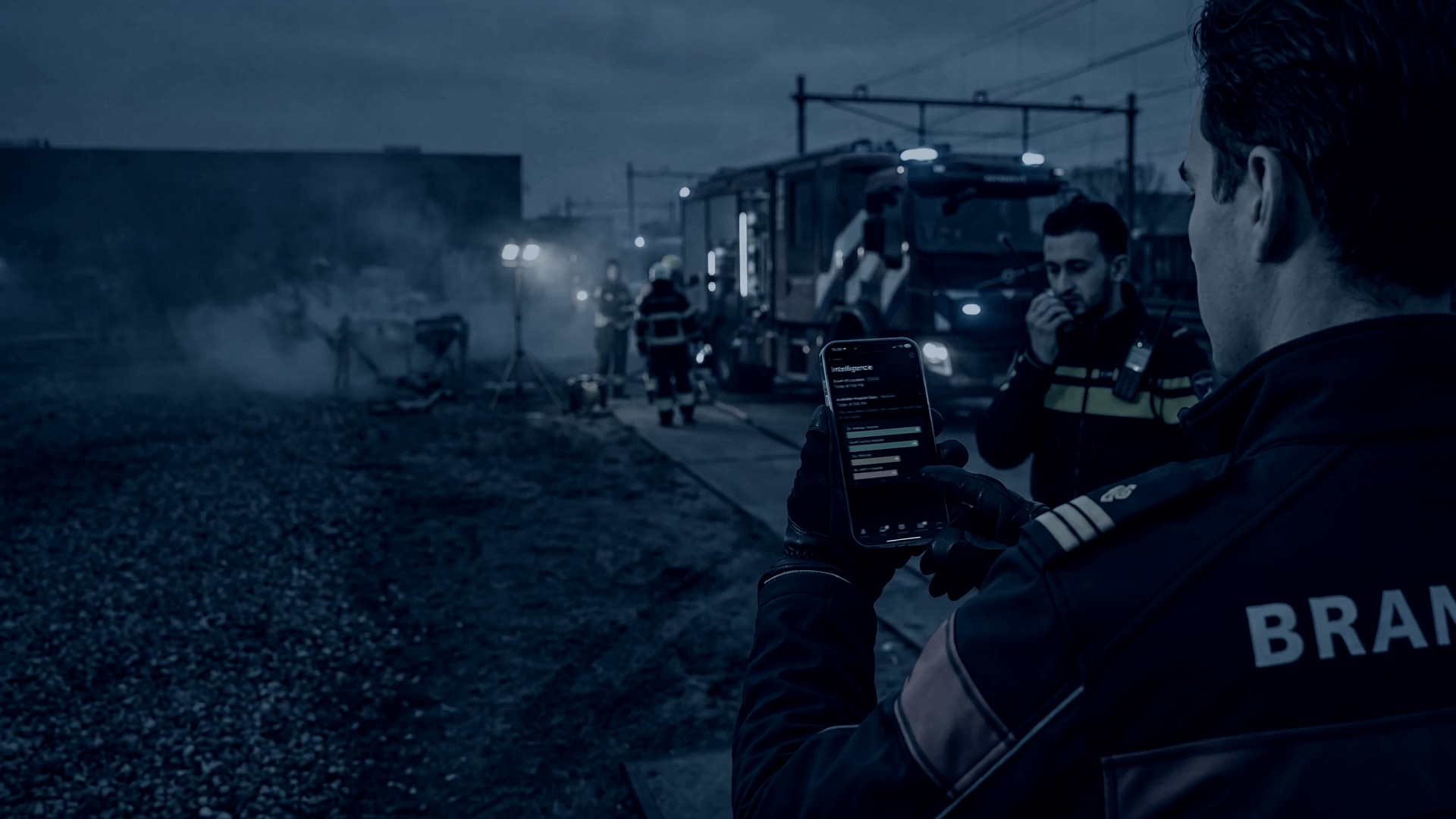 A fire officer checks live operational intelligence on his phone at an incident scene at dusk, with fire and police crews working behind him