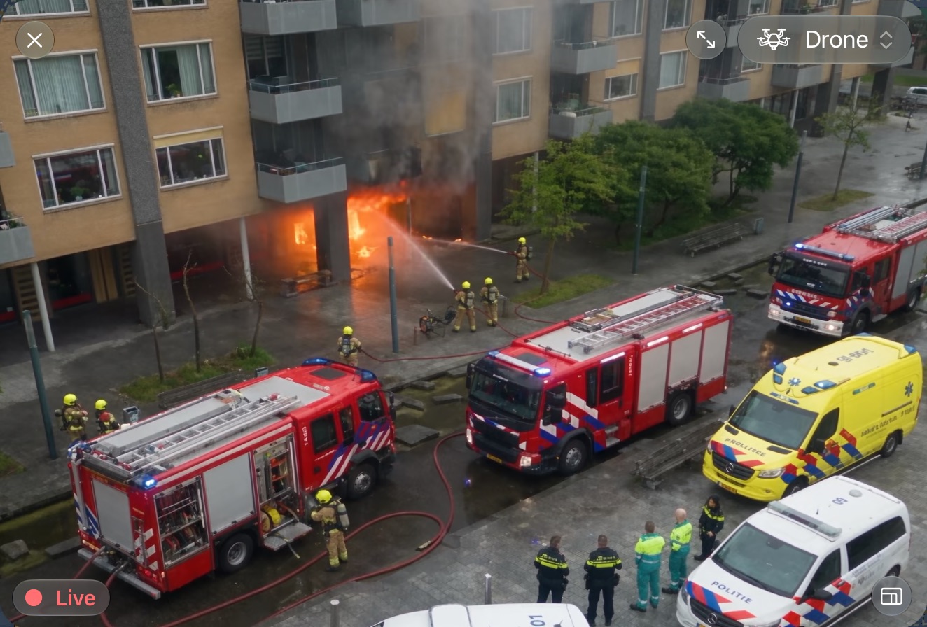 Live drone video in LIVEOP Response: fire crews tackling a fire at the foot of an apartment building, with fire engines, an ambulance and police on the street below